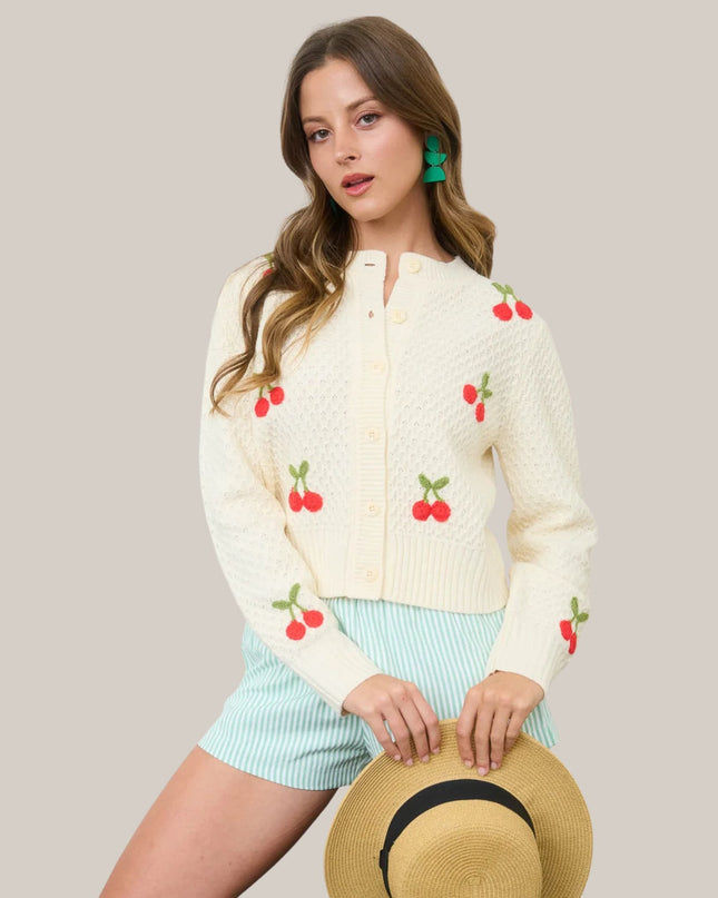 Woman wearing a cream cardigan with cherry patterns, holding a straw hat against a plain background