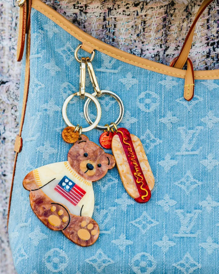 A close-up of a light blue monogram handbag with a decorative keychain charm hanging from the strap. The charm features a brown teddy bear wearing a white shirt with an American flag graphic and a red hot dog in a bun.
