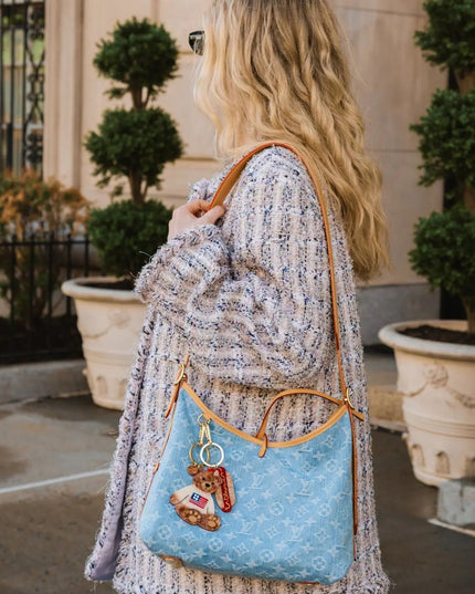 A woman wearing a light purple knit cardigan carries a light blue Louis Vuitton monogram shoulder bag with tan leather trim and a small charm, photographed outdoors in front of a building with potted plants.