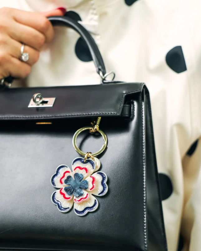 A close-up of a black leather handbag with a gold-tone turn-lock clasp and a colorful embroidered flower charm hanging from the ring. A hand with red nail polish and a ring is holding the bag handle, against a background of a cream-colored polka dot top.