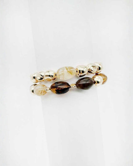 A pair of beaded stretch bracelets on a white background. One bracelet features metallic gold-toned beads, while the other features a mix of cream, clear, and dark brown marbled beads.