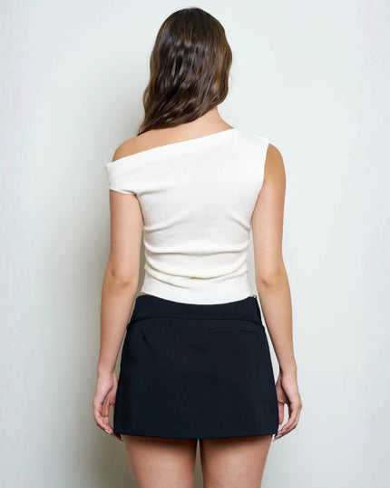 A woman shown from the back wearing a cream off-the-shoulder knit top and a black mini skirt, standing against a plain light gray wall.