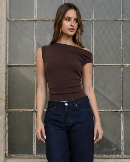 A woman modeling a dark brown off-the-shoulder ribbed knit top paired with dark blue jeans, standing in front of a large industrial window with weathered glass panes.