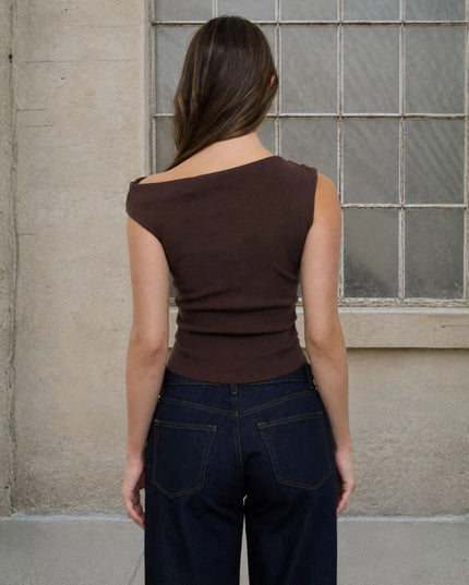A woman viewed from behind wearing a dark brown off-the-shoulder ribbed tank top and dark blue jeans, standing in front of a textured concrete wall and a window with frosted glass panes.