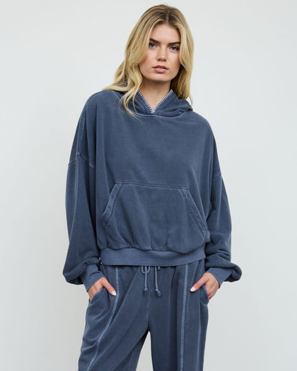 A blonde model wearing a matching dark slate blue velour loungewear set, featuring an oversized cropped hoodie with a front kangaroo pocket and matching drawstring sweatpants, set against a plain light gray studio background.
