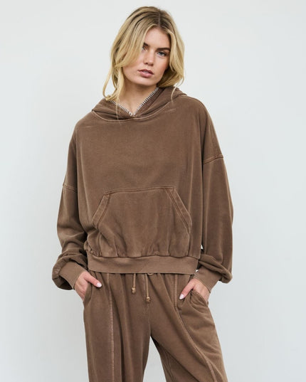 A woman modeling a matching brown lounge set consisting of an oversized pullover hoodie and matching drawstring sweatpants against a light gray studio background.