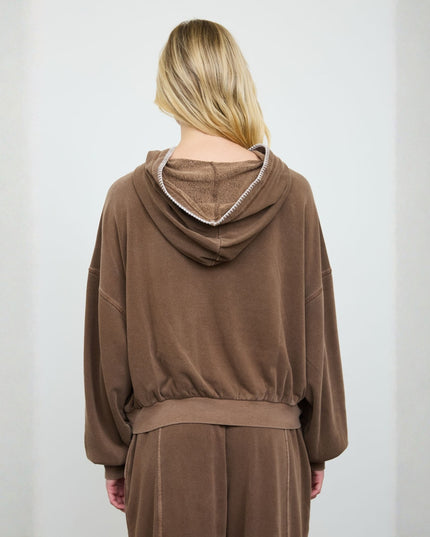 A woman viewed from behind wearing a brown hooded sweatshirt and matching pants, with a white chain detail around the hood, against a plain light gray background.