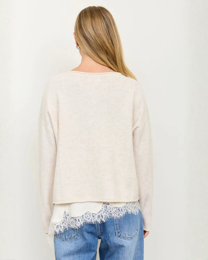 A woman viewed from the back wearing a light cream knit sweater with a lace hem detail, layered over a white top and blue jeans, against a plain off-white background.