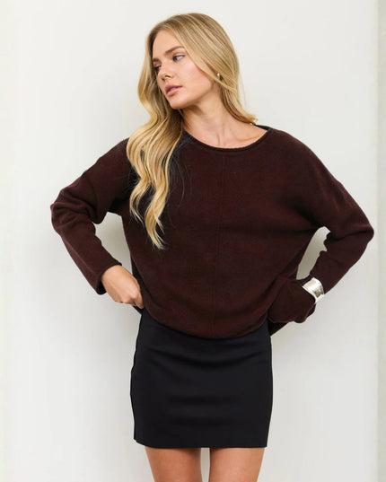 A woman modeling a dark brown long-sleeve knit sweater with a boat neckline, paired with a black mini skirt, standing against a light gray wall.