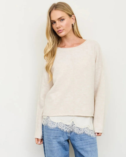 A woman modeling a cream-colored knit sweater with a wide boat neckline and long sleeves, paired with blue jeans featuring a white lace hem, against a plain off-white background.