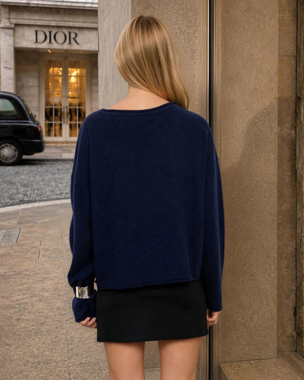 A woman viewed from behind wearing a navy blue knit sweater and a black mini skirt, standing on a city sidewalk with a Dior storefront and a black cab in the background.