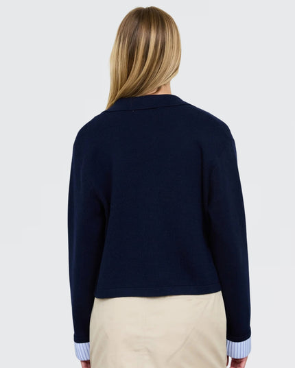A woman viewed from the back wearing a navy blue cropped knit cardigan over a light blue striped shirt and beige pants, against a plain light gray studio background.
