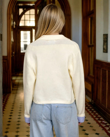 A woman viewed from behind wearing a cream-colored knit cardigan and light blue jeans, standing in a hallway with wooden doors and a patterned tile floor.