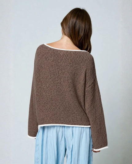 A woman viewed from behind wearing a brown textured knit sweater with white contrast trim and light blue pants, standing against a plain light gray wall.