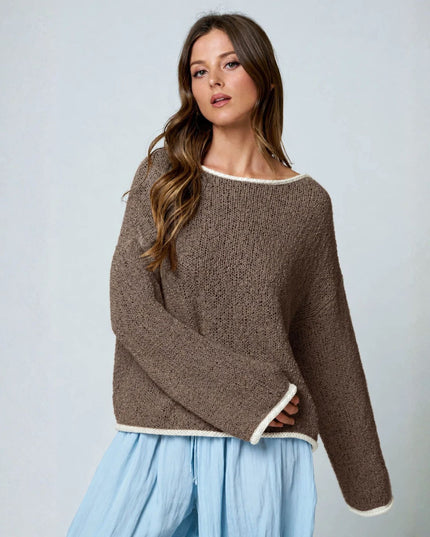 A woman wearing a brown textured knit sweater with cream trim and a wide boat neckline, paired with light blue pants, against a light gray studio background.