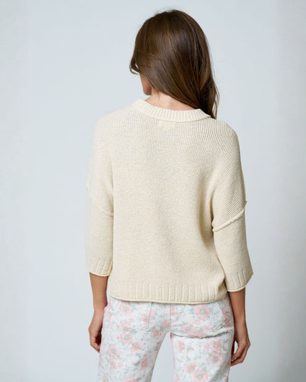 A woman viewed from behind wearing a cream-colored knit sweater and light floral pants against a plain light gray background.