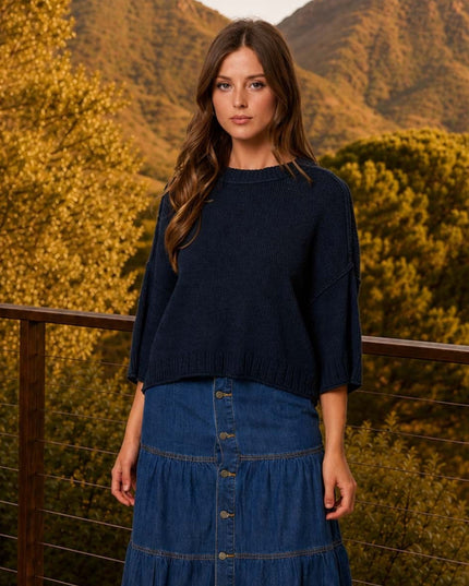 A woman stands outdoors on a balcony with mountains and trees in the background, wearing a dark navy cropped knit sweater and a blue denim tiered skirt.