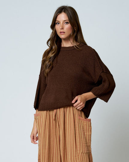 A woman modeling a dark brown knit sweater with wide three-quarter sleeves, paired with tan striped pants, against a light gray studio background.