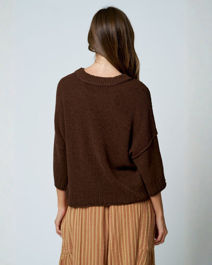 A woman shown from the back wearing a dark brown knit sweater and beige striped pants against a light gray studio background.