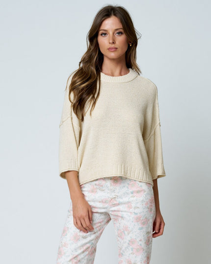 A woman modeling a cream-colored knit crewneck sweater with elbow-length sleeves, paired with light pink and white floral pants, against a solid light gray background.