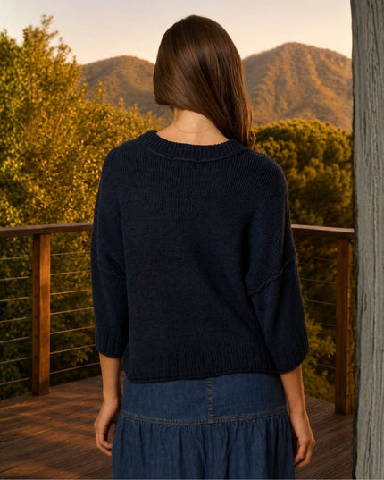 A woman viewed from behind wearing a dark navy ribbed knit sweater and a blue denim skirt, standing on a wooden deck with a scenic mountain and forest background.
