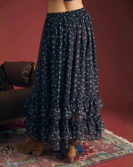 A dark navy blue maxi skirt with a small white and gold floral print, featuring multiple ruffled tiers and an elastic waistband, shown on a model in a styled room setting.