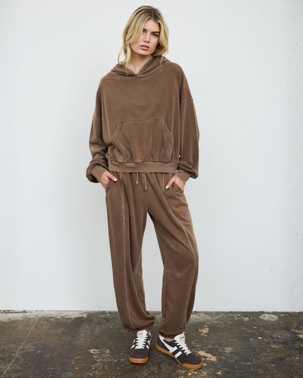 A woman modeling a matching brown lounge set consisting of a cropped pullover hoodie and relaxed drawstring sweatpants, paired with black and white sneakers, standing against a white wall.