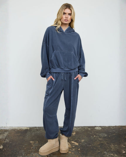 A woman modeling a matching dark slate blue lounge set consisting of a cropped pullover hoodie and relaxed sweatpants, paired with beige platform boots, standing against a white wall.