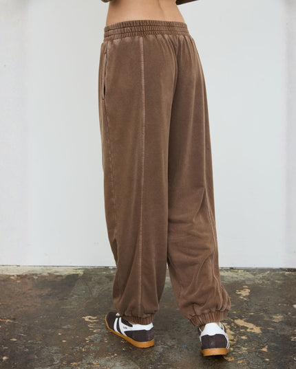 A person shown from the waist down wearing loose brown corduroy pants with an elastic waistband and elastic cuffs, paired with gray and white sneakers, standing against a white wall.