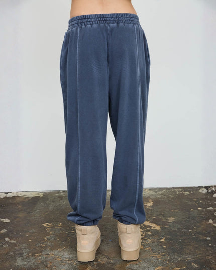 A person shown from the back wearing loose blue jogger-style pants with a gathered elastic waistband and cuffs, paired with beige platform sneakers, standing against a plain light wall and textured floor.