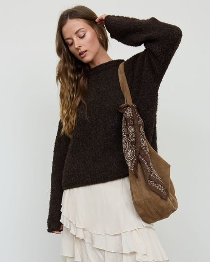 A woman wearing a dark brown fuzzy knit sweater and a white ruffled skirt, carrying a brown suede shoulder bag with a patterned bandana strap against a light gray studio background.