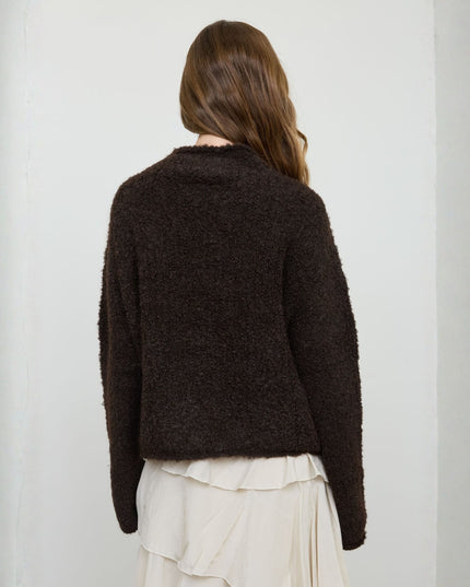 A woman viewed from the back wearing a dark brown textured boucle cardigan and a cream pleated skirt, standing against a plain light gray wall.