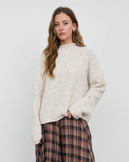 A woman modeling a chunky knit cream-colored sweater paired with a pink and brown plaid skirt, set against a plain light gray studio background.