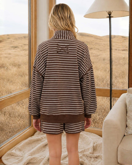 A woman stands with her back to the camera wearing a brown and beige horizontally striped lounge set consisting of a mock-neck long-sleeve top and matching shorts. She is positioned in a cozy room with large windows overlooking a dry grassy field, with a floor lamp and a cream-colored armchair visible.