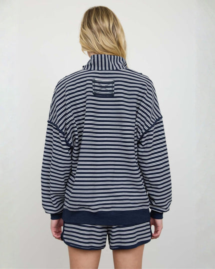 A woman viewed from behind wearing a navy and white striped long-sleeve quarter-zip jacket and matching striped shorts against a plain off-white wall.