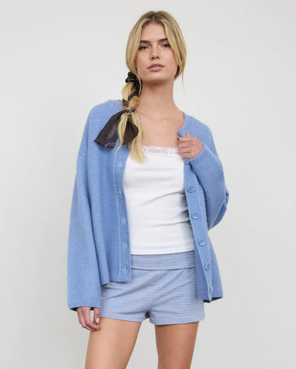 A blonde model wearing a light blue knit cardigan over a white tank top and matching light blue striped shorts, standing against a plain light gray studio background.