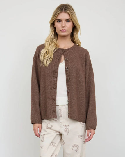 A woman modeling a brown knit button-front cardigan over a white top and cream pants with small printed motifs, photographed against a plain light gray studio background.