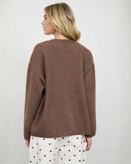 A woman shown from the back wearing a loose-fitting, heathered brown knit sweater and white pants with brown polka dots, set against a plain light gray studio background.