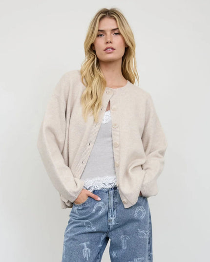 A blonde woman models a light beige knit cardigan over a gray top and blue jeans with white embroidered details, set against a plain light gray background.