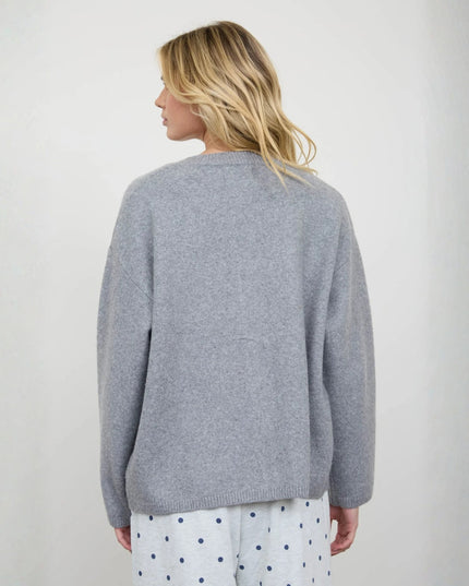 A woman viewed from the back wearing a light gray knit sweater and white pants with blue polka dots, standing against a plain light gray wall.