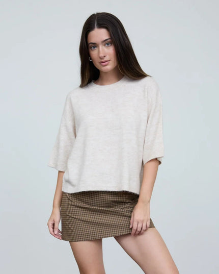 A woman modeling a cream-colored, oversized short-sleeve knit top paired with a brown and beige gingham mini skirt, set against a plain light gray studio background.