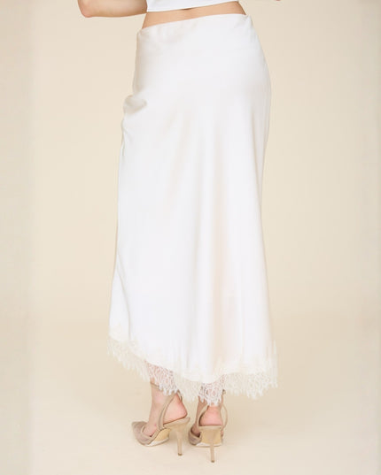 A white satin maxi skirt with a delicate lace hem, shown on a model from the back against a warm beige studio background.