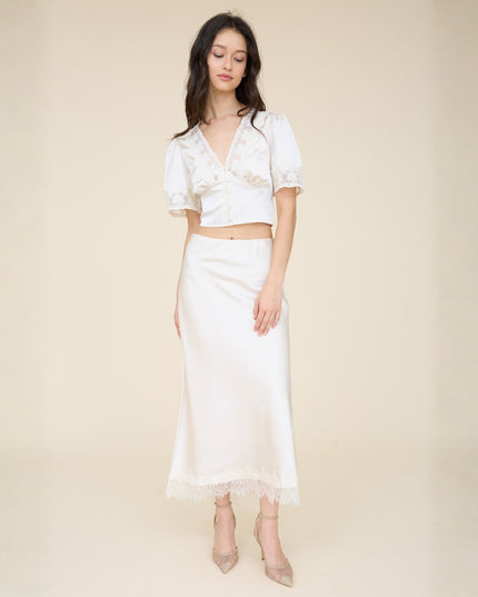 A woman modeling a two-piece white outfit consisting of a short-sleeve lace-trim crop top and a satin midi skirt with lace hem, paired with nude high heels, against a solid beige studio background.