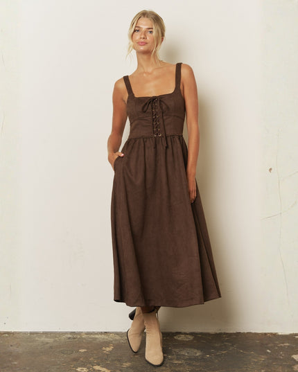 A woman modeling a brown midi dress with a lace-up corset-style bodice, square neckline, and pockets, standing against a textured off-white wall.
