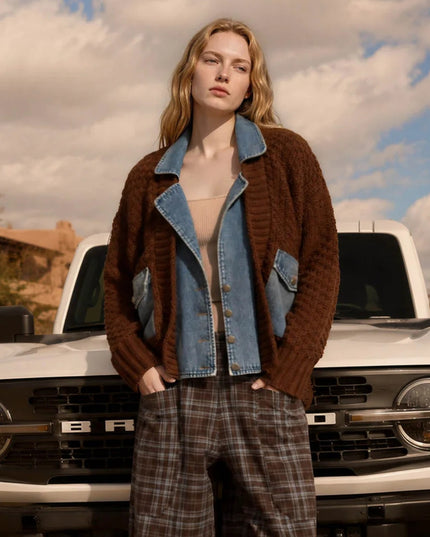 A woman stands in front of a white off-road vehicle outdoors, wearing a brown ribbed knit cardigan over a light blue denim jacket, a beige top, and brown plaid pants.