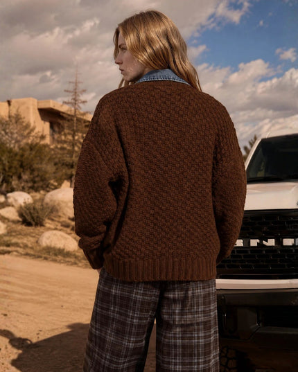 A person standing outdoors in a desert-like setting, shown from the back wearing an oversized textured brown knit sweater over a blue denim shirt and plaid pants, with a white vehicle behind them.