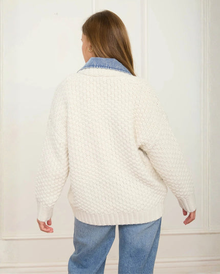 A woman viewed from behind wearing a cream cable-knit sweater over a light blue denim collared shirt, paired with blue jeans, set against a white paneled wall.