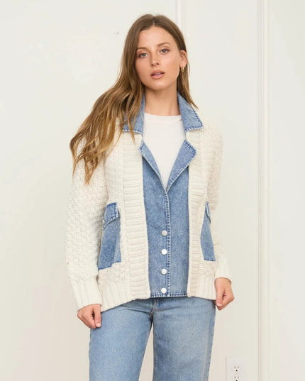 A woman modeling a cream cable-knit cardigan with light blue denim paneling and pockets, worn over a white top and light-wash jeans against a plain white wall.