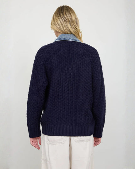 A person shown from the back wearing a dark navy textured knit sweater over a light blue collared shirt, paired with off-white pants, against a plain light gray background.