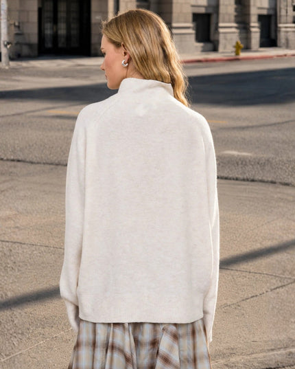 A woman stands outdoors on a city street, shown from the back, wearing a cream-colored knit mock-neck sweater and a plaid skirt.